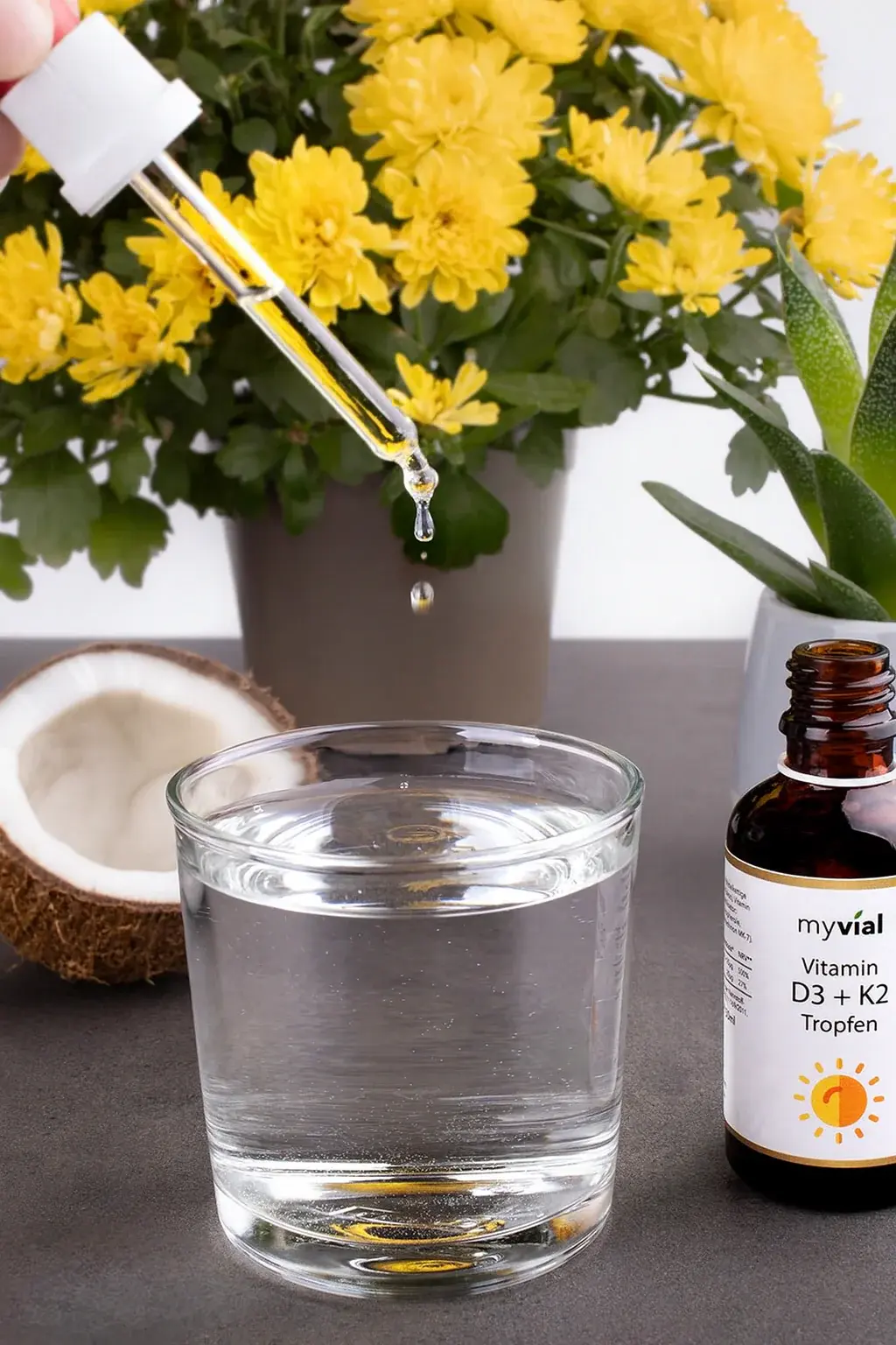 Ein Tropfen Vitamin D3 + K2 fällt in ein Glas Wasser, daneben Blumen und eine Kokosnuss